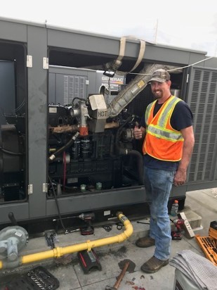 A maintenance worker in a high-visibility vest and a hard hat stands proudly next to an open industrial generator he is servicing for ACF Standby Systems.